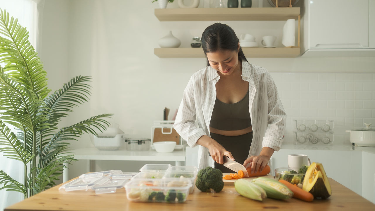 Beautiful young woman dressed in fitness wear arranging meal prep containers with healthy ingredients on a wooden countertop.